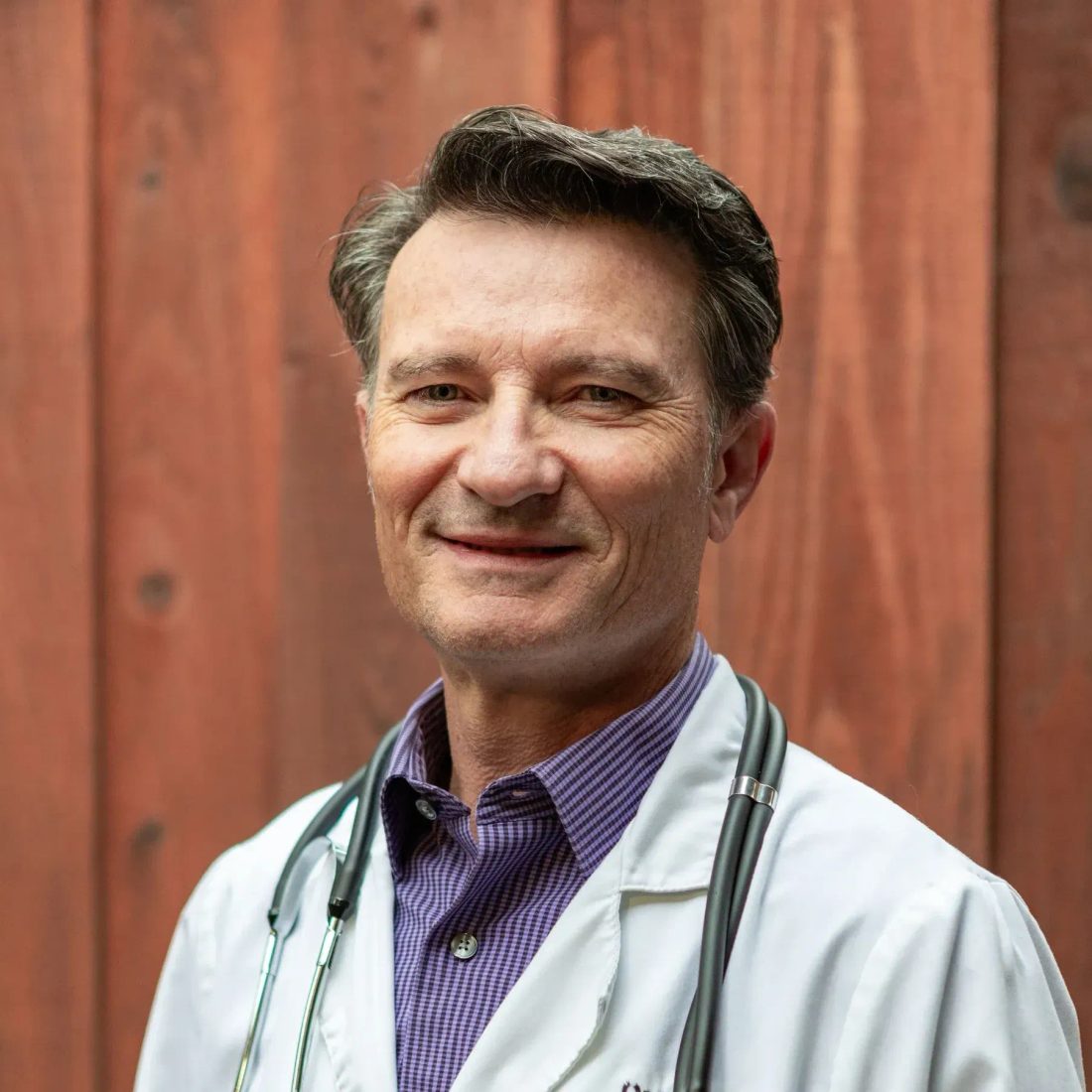 Smiling male veterinarian in white coat with stethoscope against wood wall